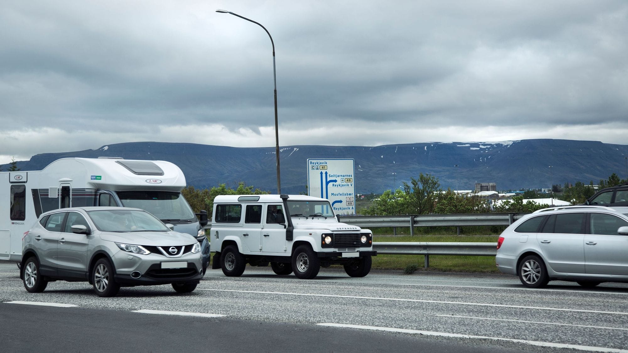 Traffic street, cars drive in two lanes, traffic sign in the middle of the picture, vegetation and houses in the background and at the very back a blue mountain